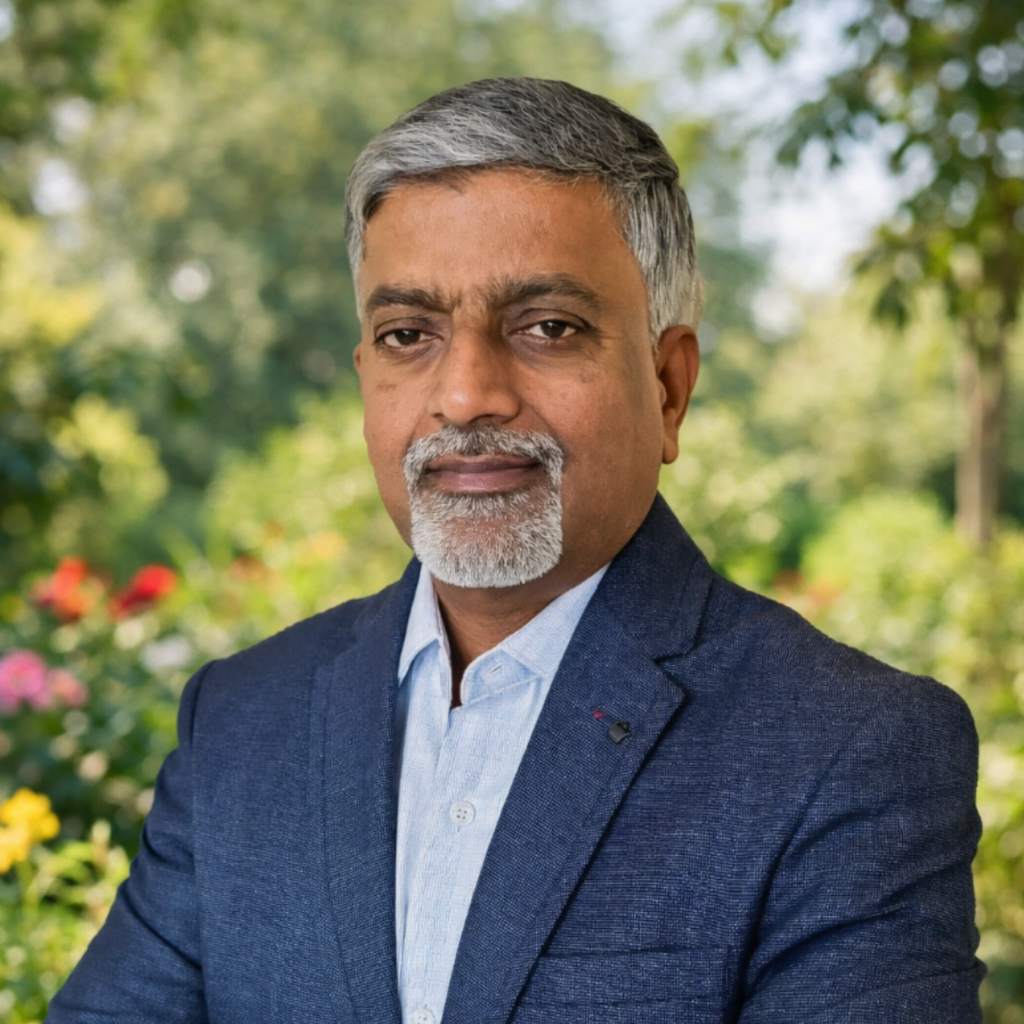 A man with grey hair and a beard wearing a blue suit jacket standing in front of a garden.