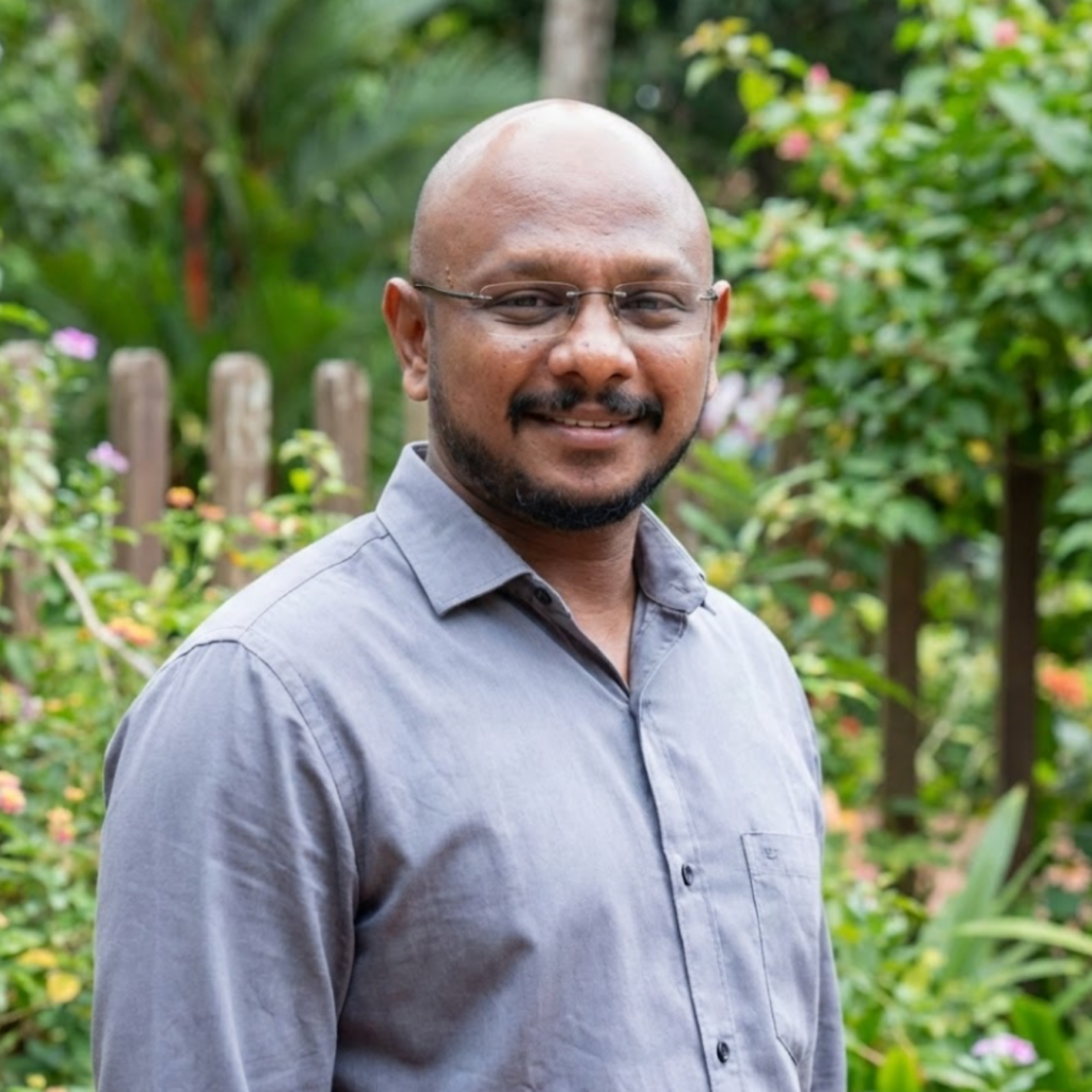 A bald man with glasses and a beard smiling in a garden with a wooden fence.